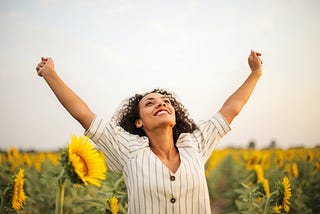 A woman in a sunflower field smiles and holds her arms skyward with positivity.