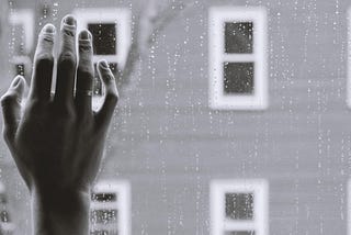 Black and white filter of a hand pressed against a window with raindrops on it.