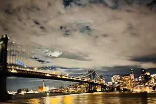 A lighted bridge is shown over water at night time with city buildings in the background