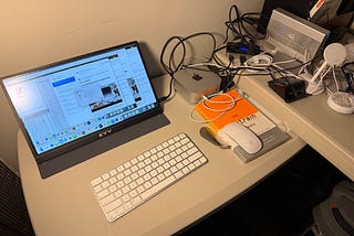 A desk setup featuring a Mac Mini connected to a KYY portable monitor displaying various application windows. The desk also has an Apple Magic Keyboard and a wired Apple mouse resting on top of a book titled “The Cluetrain Manifesto.” Behind the Mac Mini, there are several cables and connected devices, including an external hard drive and a white MagSafe phone stand. The desk is organized, though several cables are visible, creating a somewhat cluttered look on the right side.