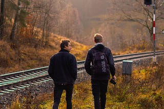 Two men in a rural environment are seen from the back, walking at the edges of a railway track while the traffic light is red