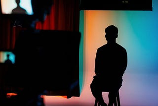 Person in sillouette sitting down in a recording studio inside Helix within the EC Stoner building. The background is colourful and in the foreground we can see a silouette of a camera and screen.