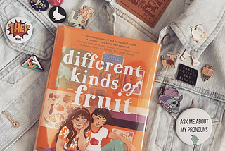 Kyle Lukoff’s middle grade novel Different Kinds of Fruit lays on a jean vest, surrounded by pronoun pins and other various enamel pins with slogans like “Babes Against the Binary” and “Gender Outlaw”