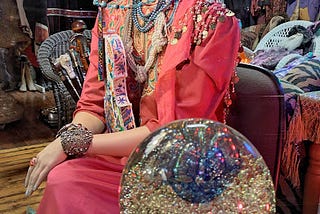A shop window mannequin, her lovely female form adorned in coral beaded and bejeweled finery against a background of magnificent costumery. In the foreground sits a glittering crystal ball filled with colors