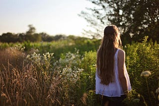 A young woman with long hair stands in alone in a meadow, her back to camera