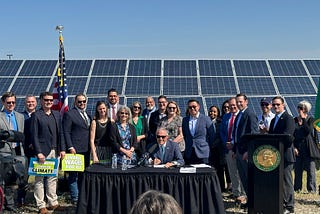 Photo of Gov. Inslee seated at a table signing a bill and a crowd of a couple dozen people standing behind him, smiling at the camera. Behind them are a large array of solar panels and bright blue sky.