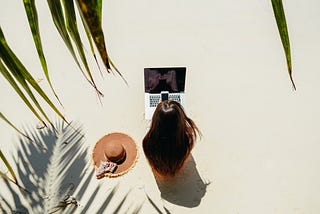 Foto tirada do ângulo de cima mostrando uma mulher sentada, de costas na praia, e trabalhando.