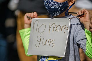 Little boy wearing a face mask and holding a handwritten sign that reads “No more guns.”