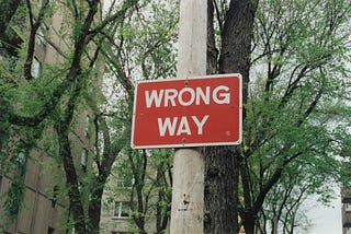 Placa vermelha escrita "wrong way", em um poste de concreto. No fundo, árvores com folhas verdes e alguns prédios.