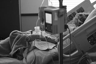In the foreground, an IV machine, unfocused. In the background, a patient lying in a hospital bed, turned towards a window.