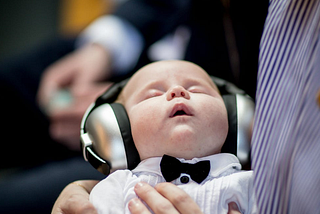 Close up photo of a baby in a white dress shirt and black bow tie with large headphones on asleep in the arms of an adult.