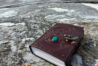 A richly embossed burgundy leather journal with a pentacle design, turquoise gemstone, and beautiful old-style clasp rests on a weathered stone surface on a beach in Norway, evoking a sense of mystery, ritual, and ancient wisdom.