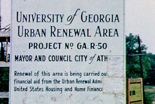 Signboard in front of construction site, reading (partly obscured): University of Georgia Urban Renewal Area Project No. GA R-50 Mayor and Council City of Ath[ens, GA] Renewal of this area is being carried ou[t with] financial aid from the Urban Renewal Admi[nistration] United States Housing and Home Financ[e Agency]