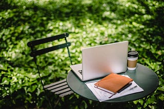 small table with black chair, laptop, journal, and coffee cup on it.