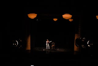 Two stage lights shine on a woman onstage. She is gesturing while performing her solo show.