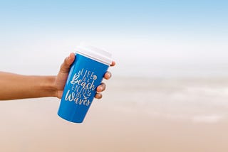 A hand holding a blue cup with words that said, “Life is a beach. Enjoy the waves.”