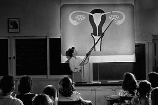 In an old black and white photo, a teacher points to a projected image of a uterus, ovaries, and fallopian tubes as students look on.