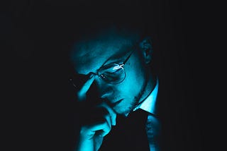 Man with glasses has his head tilted down, eyes closed and hand on the middle of his eyebrows. A blue light illuminates him.