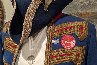 An ensemble evoking George Washington’s Continental Army hangs on a mannequin, with a blue tricorn hat embellished with a quill, and a blue coat sporting an “I Voted” sticker alongside a “Dissent” pin modeled after Justice Ruth Bader Ginsburg’s famous dissent collar.