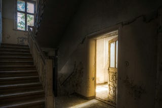 Darkened interior of abandoned asylum outside of Berlin, Germany. The interior is dark. There is a staircase to the left of the picture, with a small window at the top of the stairs. To the right of the image is a doorway, with a wooden door slightly ajar, and leading into another room. Light streams from this other room into the hallway. The rest remains in shadow.