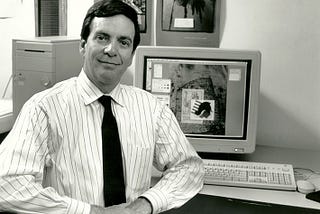 Black and white photograph of a white man with black hair sitting in front of a 1990s computer. he man is wearing a white striped button down shirt and a dark necktie. The man has a slight smile on his face. His hands are clasped in front of his body.