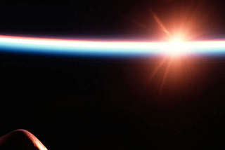 Sunrise viewed from space, with the sun flaring bright orange-white just above Earth’s curved horizon, which glows in a thin band of blue and red against the black of space. Part of a spacecraft’s dark silhouette, including a vertical fin or antenna, is visible in the lower left foreground.