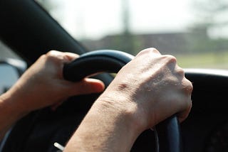 A photo of two white-skinned hands gripping a grey steering wheel.
