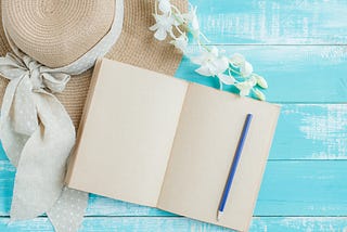 A blank notebook with a blue pencil, set next to a beach hat against a sky blue background.