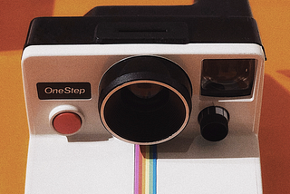 White Polaroid Land Camera with a One Step red button and rainbow running down the middle. This image has an orange background.