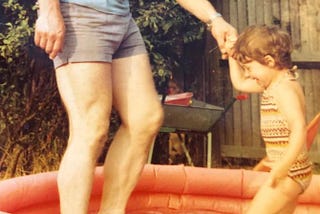 Small child in paddling pool holding her father’s hand