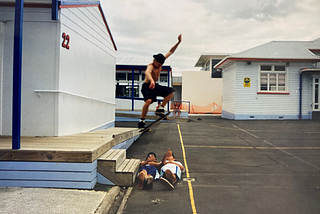 A skateboarder is captured in mid-air, he’s halfway through an olly – jumping off  two stairs and over two humans (his friends). It’s a make or break moment. He’s mid-flight directly above his two friends, who are lying down beneath him. One is throwing a shaka, the other is giving the thumbs up. Two younger kids watch the action from the distant background. They have no shoes. They wait to see if he lands it, or if he crashes into his mates.