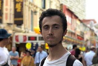 A man with long black hair looking straight at the camera. He is wearing a white shirt and a black backpack. Behind him are signs in chinese hanging from tall buildings.
