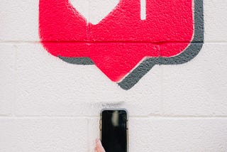 Street art of an Instagram “like” red bubble with heart on a white cement block wall, with a hand holding up a phone beneath it