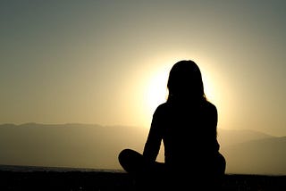 Photograph of a woman sitting cross-legged and meditating outside at sunset.