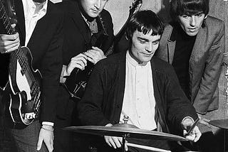 Paul McCartney, John Lennon, George Harrison and Jimmie Nicol pose for a photo. Paul is holding his bass, John is holding a guitar, Nicol is sitting with a drum set, George isn’t holding anything but is watching the drums.