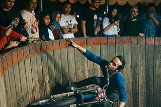 A man is riding a motorcycle around a the curve of a sheer sided wooden wall, and reaching out to receive a cash tip held out by a spectator