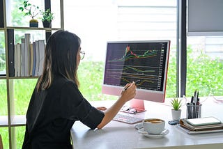 A young woman sitting in front of a large monitor showing investment performance.