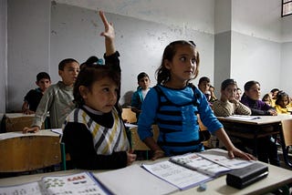 A classroom full of Syrian students, with several children visible in the foreground and others in rows behind them. In the center, a student raises their hand while another student in a blue and black striped sweater looks to the side. The students sit at wooden desks with workbooks open in front of them. The classroom has plain white walls and natural lighting coming from above. The children appear engaged and attentive, dressed in casual clothing including striped sweaters and solid-colored s