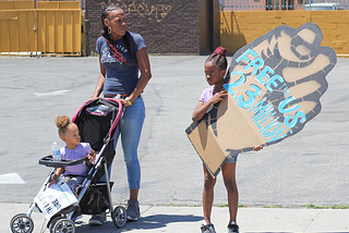 Black mother pushing stroller with toddler, standing next to a child with a fist that says, “Free us 23 million”