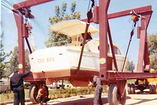 White cabin-cruiser boat that says “Cee Bee” on the back — being hoisted onto a trailer. A man stands at the back of the boat and trailer while another man is working on the right side and another man is squatting down underneath.