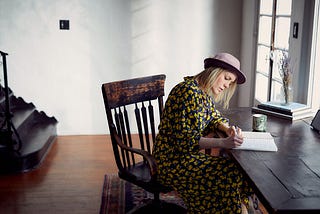 Lady wearing yellow and brown floral dress and a hat sitting at a desk in an antique wooden chair, writing in a journal. Coffee cup and books on the desk.