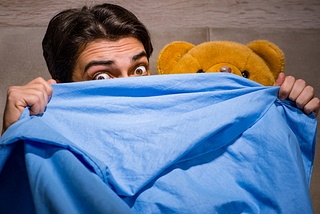 Man hidding under a blue blanket with his brown teddy bear