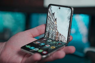 A hand holding a foldable smartphone with the home screen displayed. There is a picture of houses on a street as the background image on the phone.