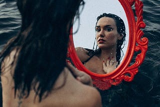 A beautiful woman sitting on the water looking at her reflection at a red mirror.
