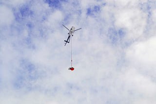 na imagem, um céu azul com nuvens e no centro, um helicóptero com uma corda e na ponta um saco laranja pendurado