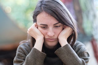 A woman, depressed and defeated, sits with her head in her hands while she looks down at the floor.