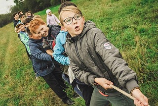 A line of young kids tugging on a rope in a green field.