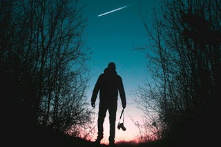 A silhouette of a man walking through a quiet forest, looking up at a bright white comet streaking across the twilight sky.