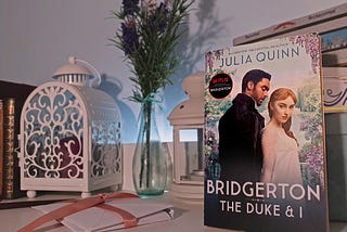The first book of the series can be seen on the right side of the picture, with the two actors from the TV show on the front of the book. Simon is facing backwards but looking at Daphne, while Daphne is looking at the objective/reader. A stack of vintage boxes keep the book upright. Two lanterns can be found in the background, along with old watermarked books on one side and purple and white flowers between the lanterns. A stack of envelopes is at the front.