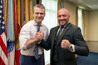 Pete Hegseth in a shirt and tie with one sleeve rolled up showing a We The People tattoo. He has locked fists in a friendly way with Conor McGregor, who is wearing a suit, tie and jacket, showing a gold watch.